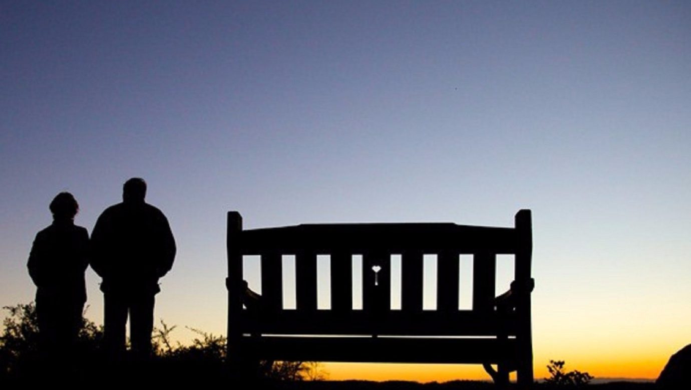 Two people stand next to a park bench looking at the view below them, at sunset