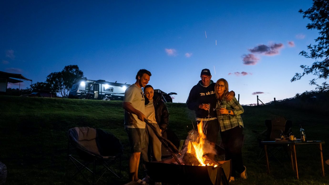 A group of friends around a campfire with their campsite in the background.