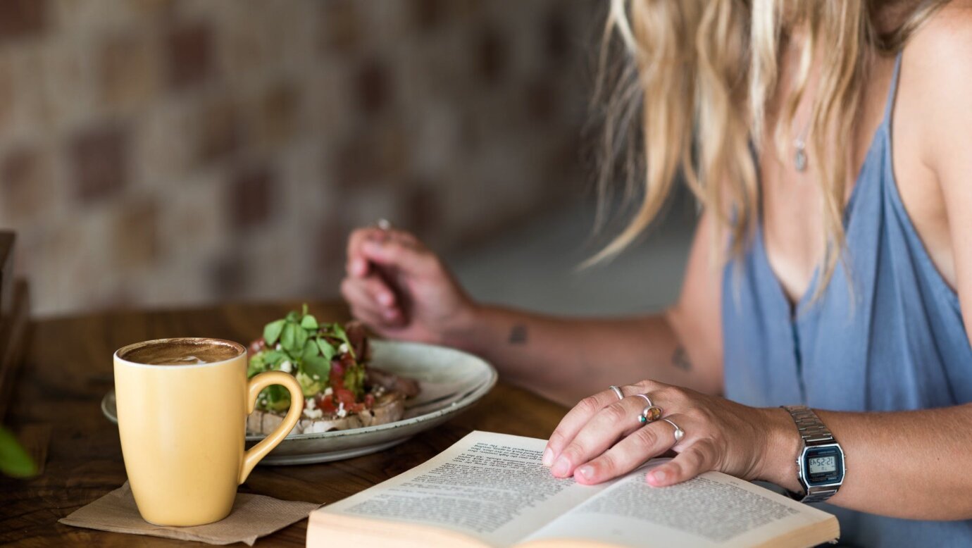 Person with long blonde hair reading a book while eating breakfast and drinking a coffee in mug