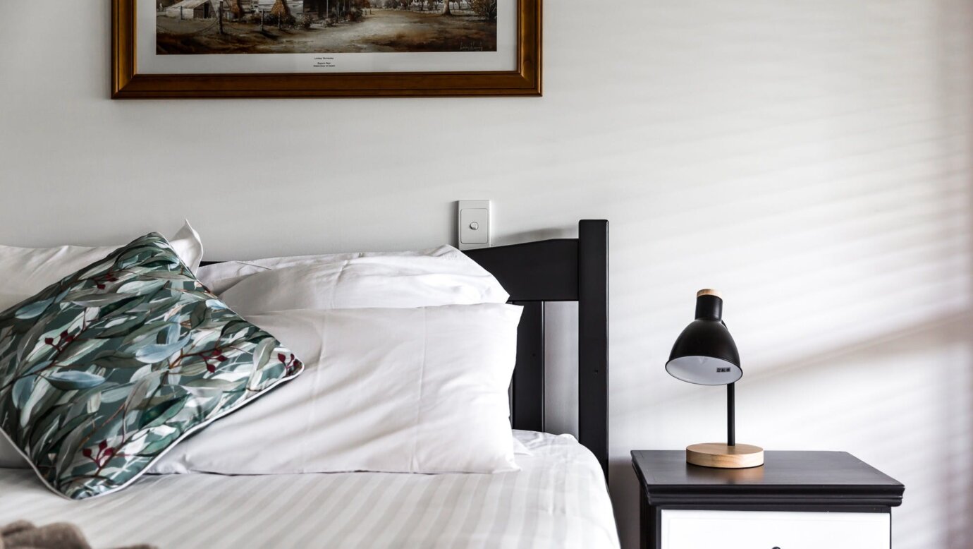 Bed with black wooden frame. White linen. Black bedside table with black lamp. Painting in backgroun