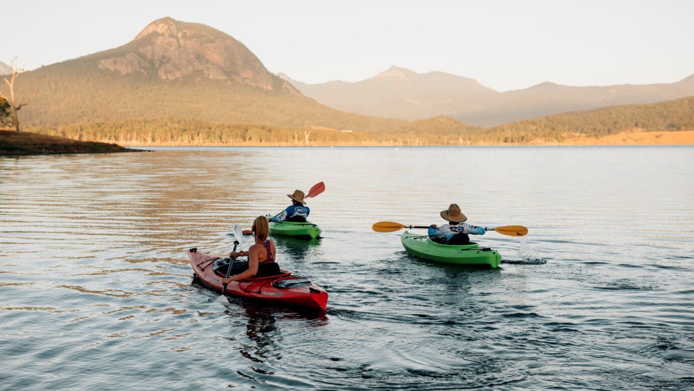 Three people kayak across a lake towards a distant mountain range