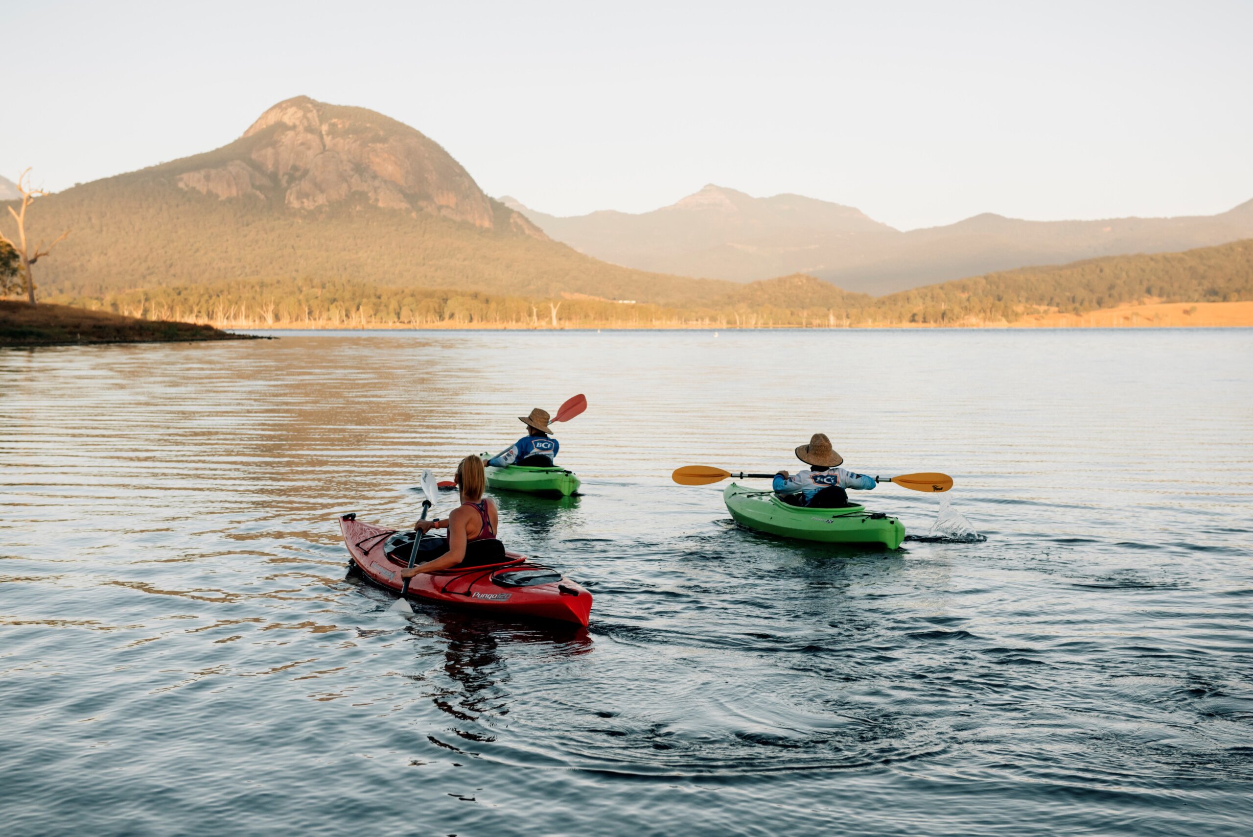 Three kayakers paddle across Lake Moogerah, with a range of mountains in the distance