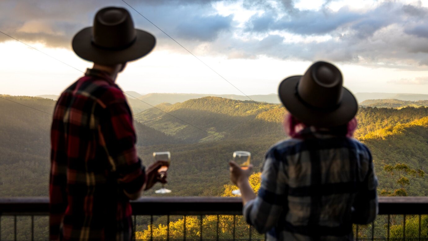 A couple stand at a balcony railing with a glass of wine in their hand looking at the valley below
