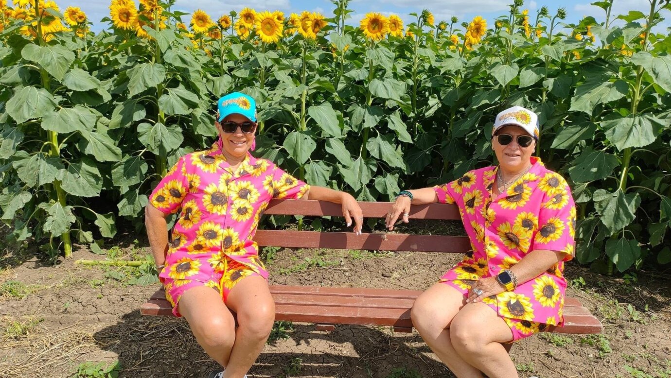 Two ladies dressed in sunflower shorts and shirts  are seated in front of the crop.