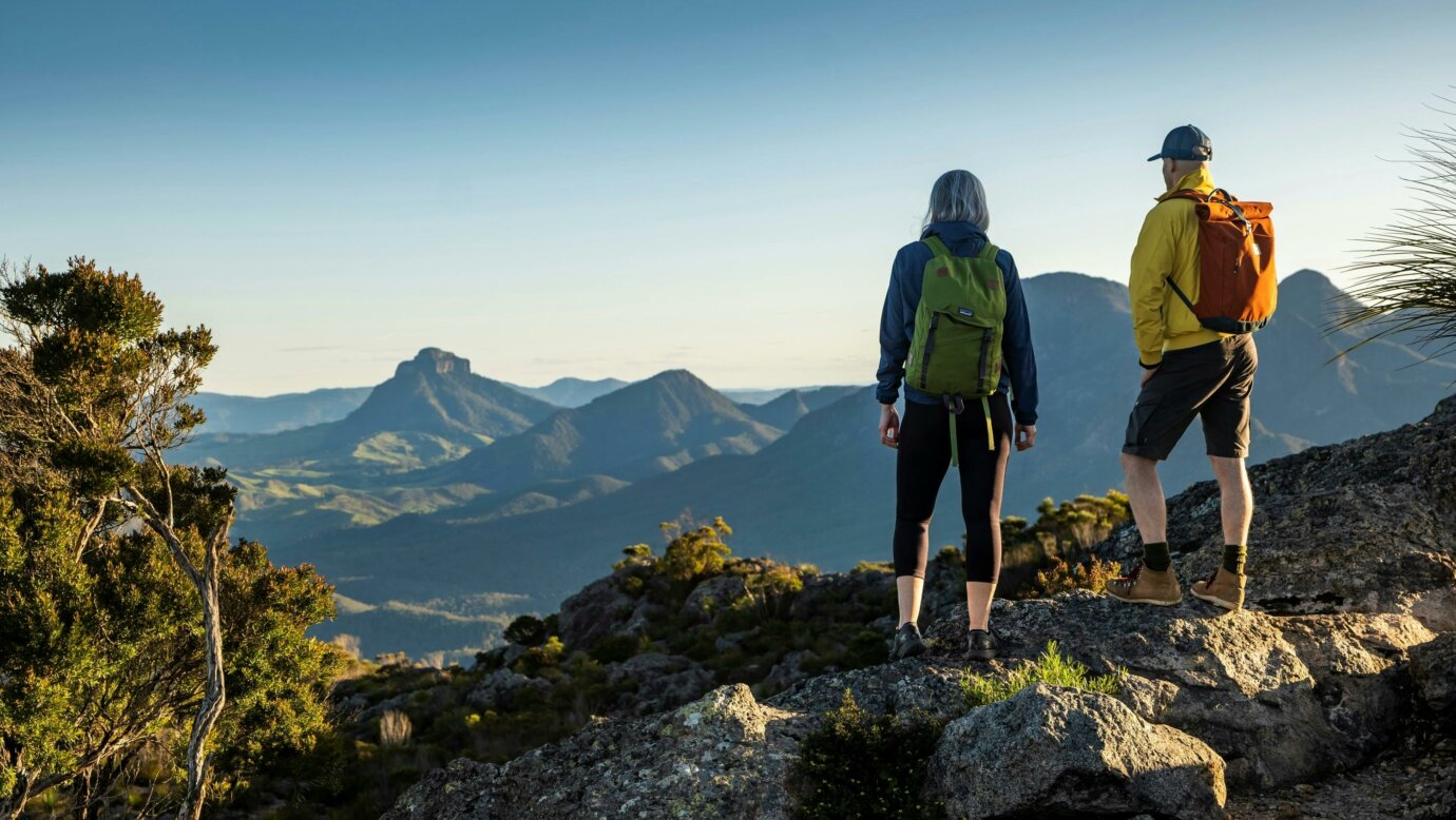 Two hikers stand on the top of a mountain, looking towards another mountain