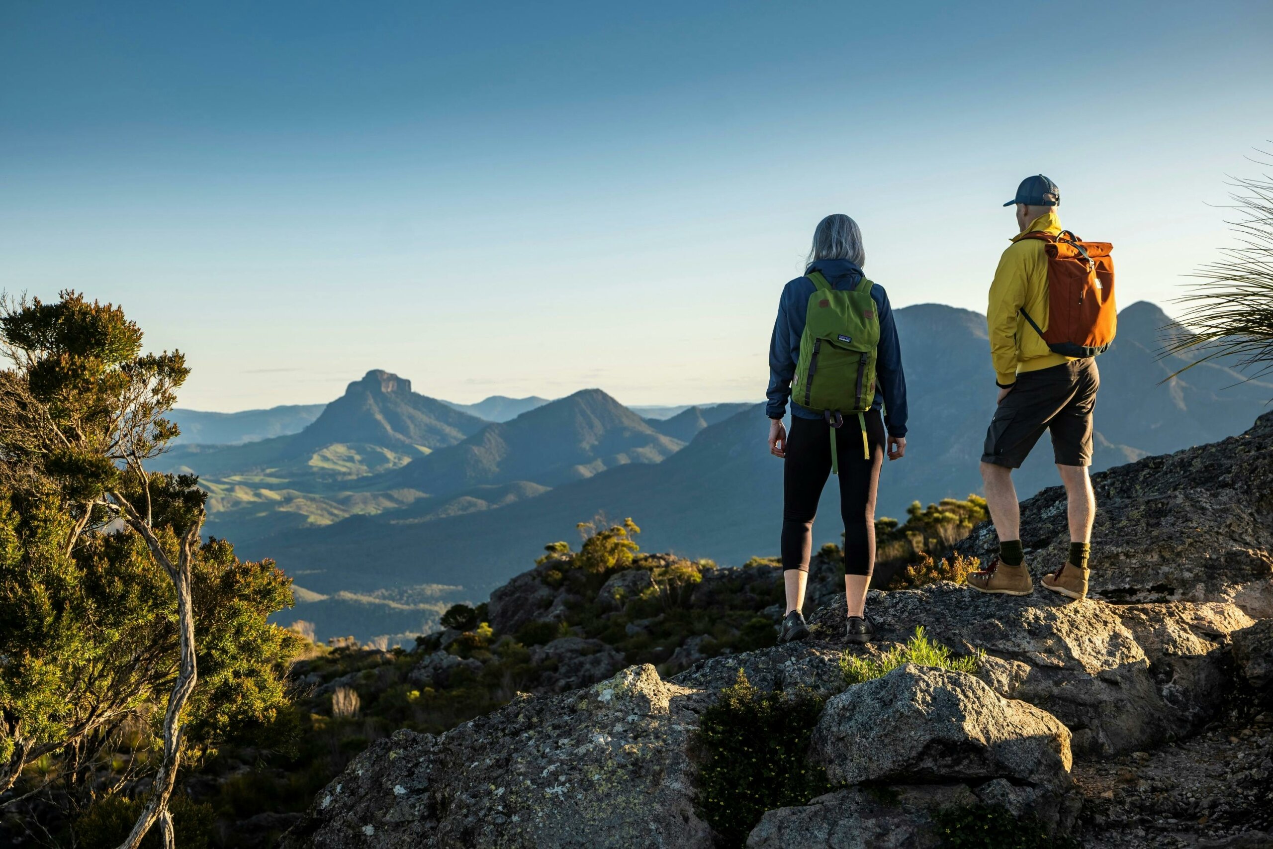 Two hikers stand on the top of a mountain, looking towards another mountain