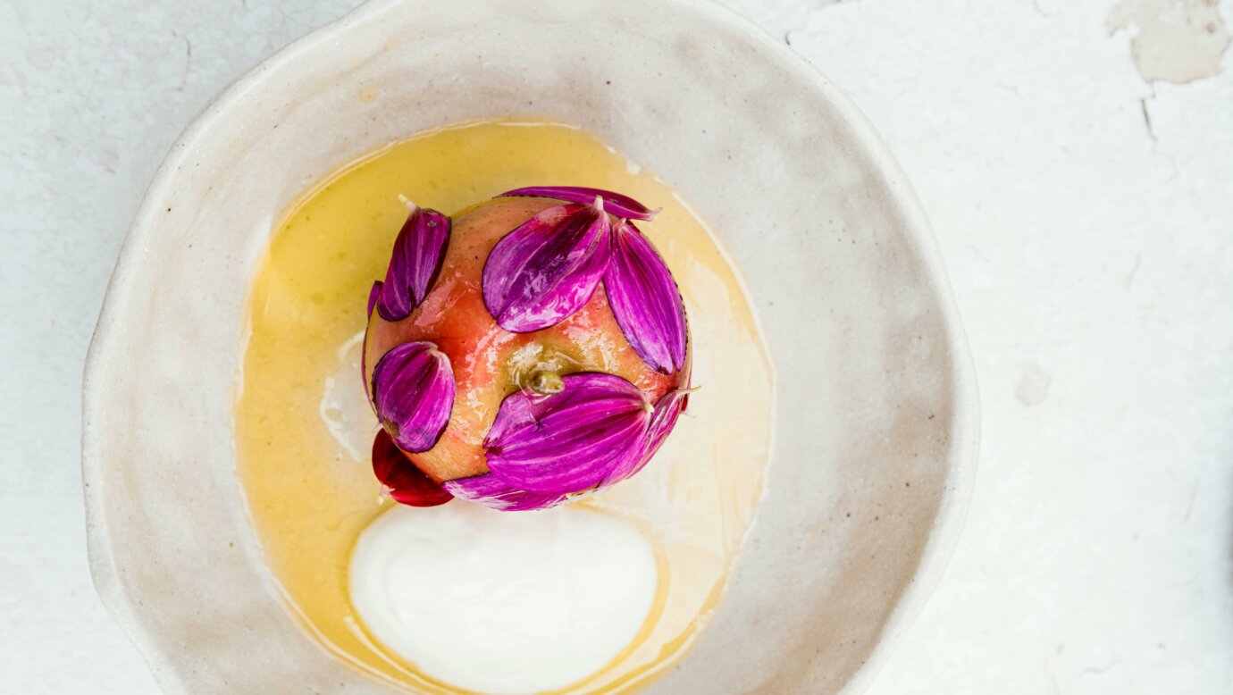 A whole slow cooked apple is served on white plate with lamb fat caramel and flower petals over it