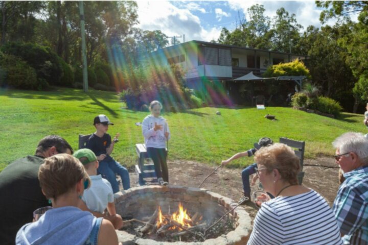 family toasting marshmallows around a firepit in the sunshine  in front  of  house and lush gardens