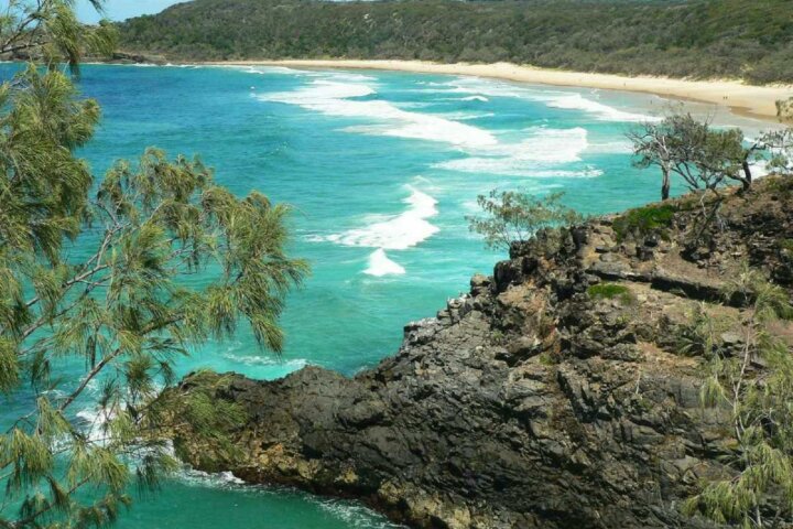 A blue water beach cove with a long stretch of white sand surrounded by hinterland and rocky outcrop